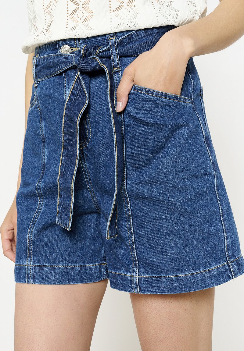 Denim šorti vidējā zilā krāsā ar sasietu jostas daļu, priekšējām kabatām un kontrastējošiem šuvju ieliktņiem.