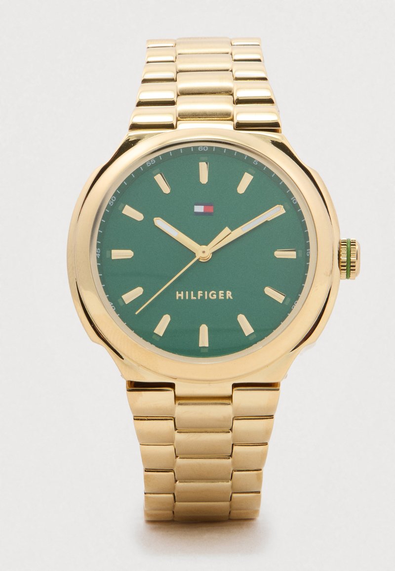 Złoty zegarek na rękę z zieloną tarczą, złotymi znacznikami godzin i wskazówkami, logo Tommy Hilfiger oraz bransoletą w formie ogniw.