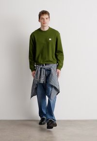 Sweatshirt verde com pequeno logo, jeans azuis e camisa de flanela cinza xadrez atada à cintura. Sapatos pretos, de pé sobre um fundo neutro.