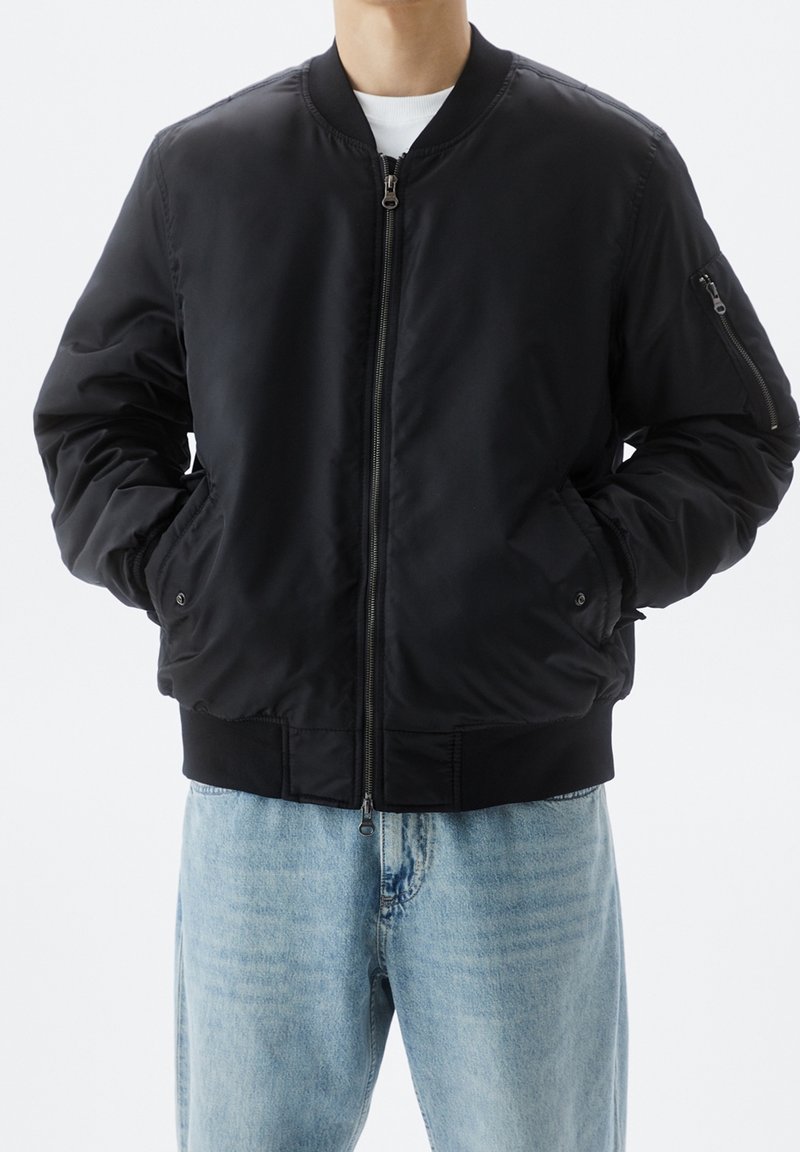 Blouson aviateur noir avec fermeture éclair devant, poignets et ourlet côtelés, doté de poches latérales et d'une poche zippée sur le bras. Porté avec un jean bleu clair.