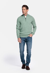 Lichtgroene sweater met kwartrits in een gebreide structuur, gecombineerd met blauwe jeans en bruine enkellaarsjes, tegen een witte achtergrond.