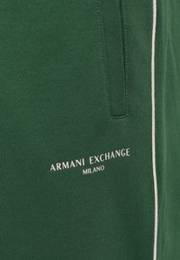Zelené textúrované joggery s bielym bočným pruhom a vytlačeným logom "ARMANI EXCHANGE MILANO" v bielej farbe blízko spodnej časti.