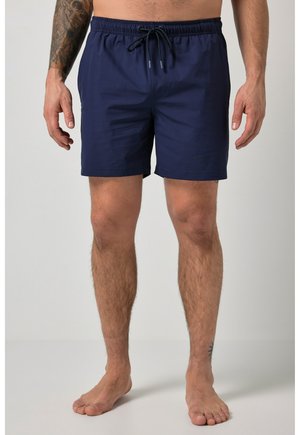 Mann, der marineblaue Badeshorts mit Kordelzug trägt und barfuß vor hellem, schlichtem Hintergrund steht.