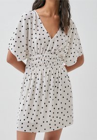 Lyhythihainen mekko kevyestä kankaasta, V-pääntiellä, rypytetystä vyötäröstä ja mustista pilkuista valkoisella taustalla.