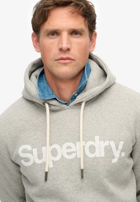 Hoodie en coton gris avec une poche kangourou à l'avant, logo "Superdry." blanc, cordons de serrage et un col de chemise en chambray visible en dessous.