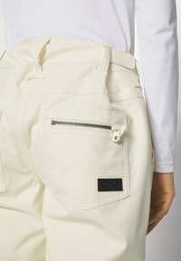 Kremfargede cargo-bukser med baklommer; en av dem har glidelås. Inkluderer en svart Roxy-logo-patch på høyre lomme.