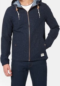 Mörkblå zip-jacka med fläckigt mönster, snörning i huvan och beige detaljer. Har två sidofickor och en liten etikett på fållen.