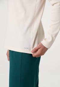 Camicia a maniche lunghe beige chiaro con texture a coste e spacco laterale, con logo "LYB" blu sull'orlo. Abbinata a pantaloni verdi.