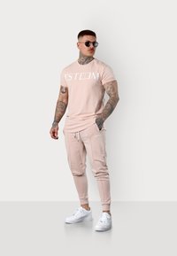 Helles pinkes Baumwoll-T-Shirt mit weißem Logo, kombiniert mit passenden Jogginghosen und weißen Sneakers. Das Model trägt dunkle Sonnenbrillen.