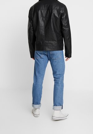 Mann, der hochgekrempelte blaue Jeans, weiße High-Top-Sneaker und eine schwarze Lederjacke trägt, steht mit dem Rücken zum Betrachter vor einfarbigem Hintergrund.