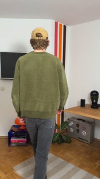 Zeleni pleteni pulover z okroglim izrezom, ohlapnim krojem in rebrasto teksturo. Kombiniran s sivimi kavbojkami, viden od zadaj v sobnem ambientu.