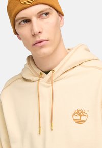 Sudadera beige con un logo de árbol dorado, con costuras en contraste y cordones dorados con puntas metálicas, llevada con un gorro de punto mostaza.