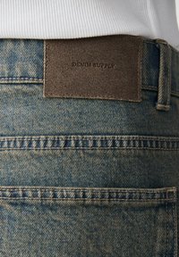Gros plan sur la ceinture arrière et la poche d'un jean en denim bleu avec un patch en cuir marron portant l'inscription "DENIM SUPPLY".