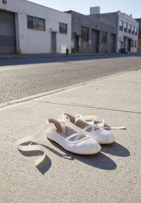 Paire de chaussons de ballet blancs avec rubans posés sur un trottoir ensoleillé, bâtiments industriels et rue vide à l’arrière-plan.