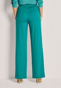 Vrouw in teal wijde broek en beige hoge hakken, staand tegen een effen lichte achtergrond.