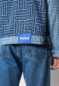 Kurtka dżinsowa z niebieskim i jasnoniebieskim wzorem logo HUGO, z niebieską metką na dole i metalowymi dodatkami. Połączona z klasycznymi niebieskimi dżinsami.
