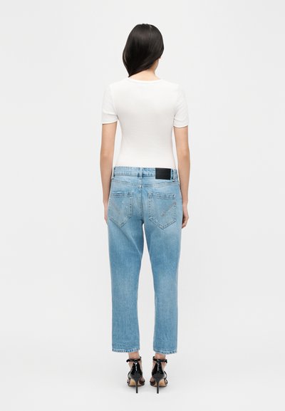 Γυναίκα, εμφανιζόμενη από πίσω, φοράει εφαρμοστό λευκό τοπ με κοντά μανίκια, ανοιχτόχρωμα μπλε τζιν ψηλόμεσα, και μαύρα σανδάλια με ψηλό τακούνι σε λευκό φόντο.