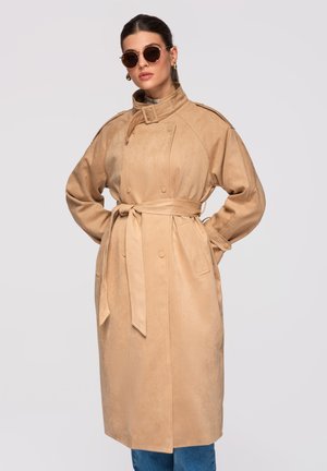 Frau trägt beigen Trenchcoat mit Gürtel, runde Sonnenbrille, Hände in die Hüften gestützt vor hellem, einfarbigem Hintergrund.