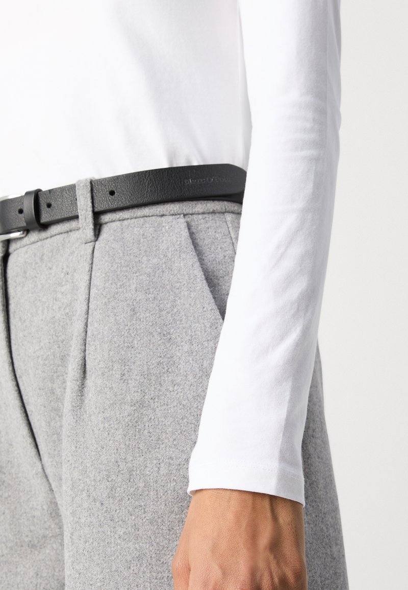 Pantaloni in lana grigia con pieghe frontali, abbinati a una cintura in pelle nera. La camicia bianca a maniche lunghe presenta una texture liscia.