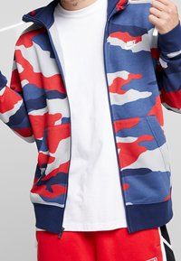 Röd, vit och blå camo zip-up jacka med en slät textur och en Nike-logotyp på vänster bröst. Har kloka fickor och ribbade muddarna.