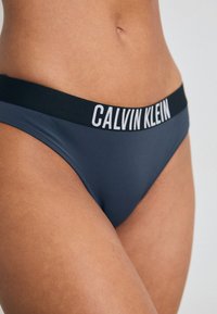 Bikini hlačke Calvin Klein temno sive barve z črnim elastičnim pasom, na katerem je v beli barvi prikazan logotip blagovne znamke. Gladka tekstura in prilegajoča se zasnova.