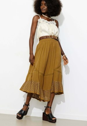 Femme portant un haut blanc sans manches orné de dentelle, une jupe midi moutarde avec un ourlet à motif, une ceinture marron, des sandales à plateforme noires et des bijoux en perles.