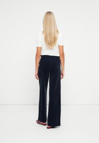 Vrouw met lang blond haar, gekleed in een witte top met korte mouwen, hoogsluitende marineblauwe brede broek en rode platte schoenen, die met haar rug naar de camera staat.
