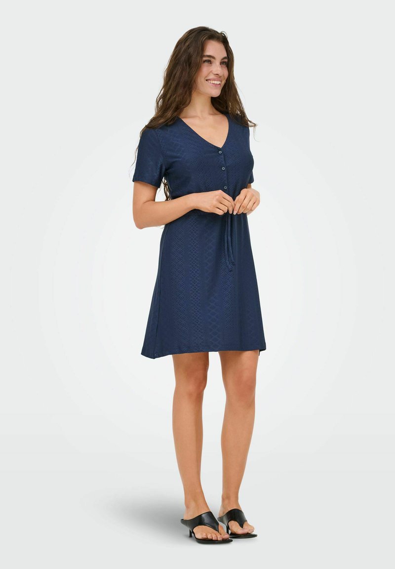 Jeune femme souriante, portant une robe bleu marine boutonnée à manches courtes et des talons ouverts noirs, debout devant un fond uni.