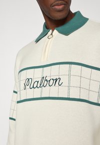 Pull polo en tricot crème avec col vert. Comprend une fermeture éclair avant et un logo "Malbon" brodé sur un motif en grille.