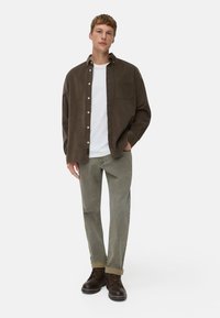 Ciemnobrązowa koszula z cordurowego materiału z guzikami, na białym t-shircie, w połączeniu z jasnoskalnymi spodniami i brązowymi butami na sznurówki, stojąca na białym tle.