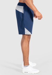 Pantalones cortos deportivos de color azul marino con un panel lateral gris, cinturilla elástica y un bolsillo con cremallera. Se llevan con zapatillas blancas.