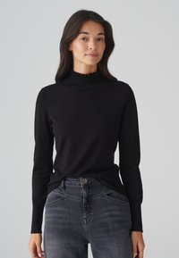 Schwarzer Langarmpullover mit hohem, rüschendem Ausschnitt, aus glattem Stoff; getragen mit dunkelgrauen Jeans mit dezenter Verblassung.