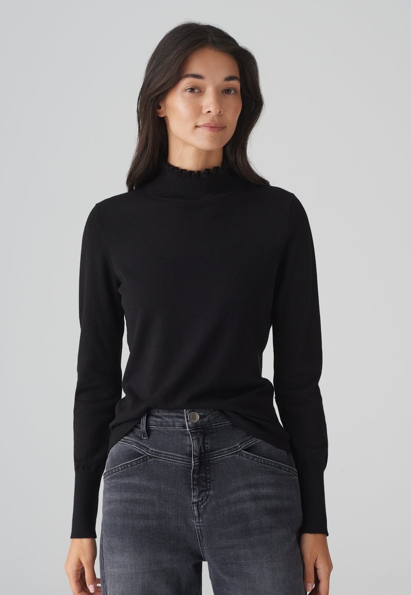 Schwarzer Langarmpullover mit hohem, rüschendem Ausschnitt, aus glattem Stoff; getragen mit dunkelgrauen Jeans mit dezenter Verblassung.