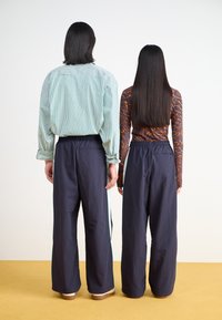 Δύο άτομα που κοιτούν προς την αντίθετη κατεύθυνση, το ένα φοράει ένα ριγέ πράσινο πουκάμισο και ναυτικές παντελόνες, ενώ το άλλο φοράει μια μπλούζα με πορτοκαλί σχέδια και ναυτικές παντελόνες με ελαστικές μανσέτες.