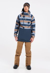 Veste de neige avec un design à motifs marron et bleu, dotée d'une capuche et d'une poche kangourou; associée à un pantalon de neige marron et des bottes noires.
