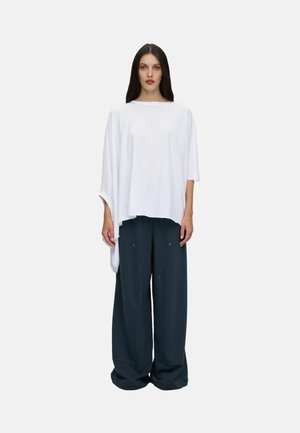 Femme aux longs cheveux foncés, portant un haut blanc asymétrique ample et un pantalon large bleu foncé, debout devant un fond uni.