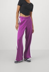 Fioletowe spodnie sportowe z białymi paskami po bokach, elastycznym pasem i luźnym krojem. W zestawie z czarnym dopasowanym topem typu crop i sportowym obuwiem.