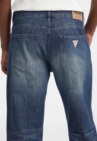 Donkerblauwe denim jeans met een vervaagde afwerking, voorzien van een tan leren label aan de achterkant en een klein rood driehoeklogo op de zak.