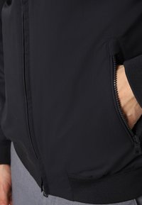 Giacca bomber nera con una texture liscia, dotata di tasca con zip e polsini a costine. Il materiale è leggero e resistente.