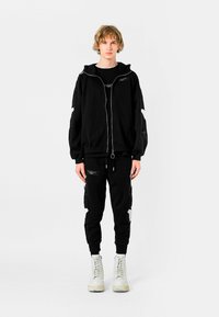 Joven de pie, vistiendo una sudadera negra con capucha y cremallera, y pantalones a juego, con botas blancas, sobre un fondo blanco liso.