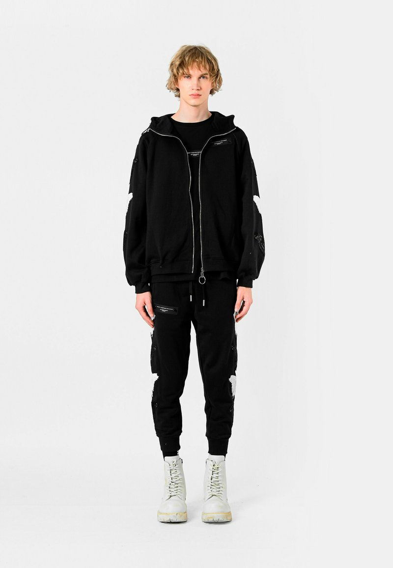 Joven de pie, vistiendo una sudadera negra con capucha y cremallera, y pantalones a juego, con botas blancas, sobre un fondo blanco liso.