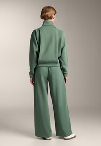 Tuta in tessuto morbido verde con colletto alto, maniche raglan e pantaloni a gamba larga. Sneakers bianche con suole marroni completano l'outfit.