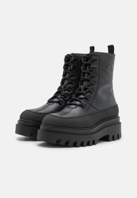 Bottes noires à cheville avec une semelle épaisse, un revêtement texturé et une pointe renforcée. Équipées d'un système de laçage et d'un détail de logo sur le côté.