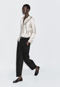 Lichtbeige satijnen shirt, zwarte corduroy broek en zwarte loafers. Losse pasvorm met een kraag en een voorzak. Gladde textuur, minimalistisch ontwerp.