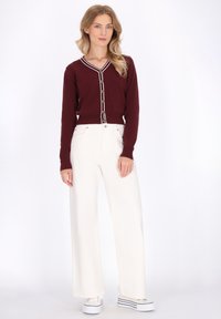 Woman wearing a burgundy buttoned cardigan with white trim, white wide-leg pants, and white platform sneakers against a plain white background.