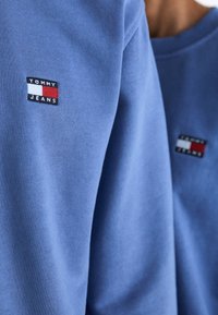Sudadera azul con tejido texturizado. Presenta un pequeño logotipo bordado de "TOMMY JEANS" en el pecho. Cuello redondo y corte relajado.