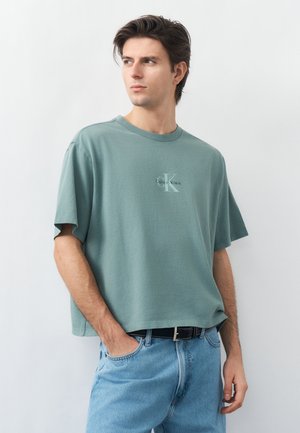 Νεαρός άνδρας με καστανά μαλλιά που φοράει μια φαρδιά πράσινη μπλούζα Calvin Klein και ανοιχτόχρωμο μπλε τζιν, στέκεται μπροστά σε ένα απλό λευκό φόντο.