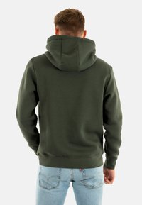 Sudadera verde oscuro con una gran capucha suave, puños y bajo acanalados. La tela parece lisa con un ajuste casual, combinada con jeans azul claro.