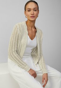Crème-kleurige gebreide cardigan met een gestructureerde, open weving. Voorzien van een ronde halslijn en geribbelde mouwen, gedragen met een wit shirt en lichte broek.