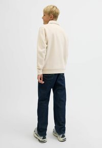 Ljusbeige fleecejacka med ståkrage, tillsammans med mörkblå denimbyxor, bärs med grå och svarta sneakers. Bakifrån.